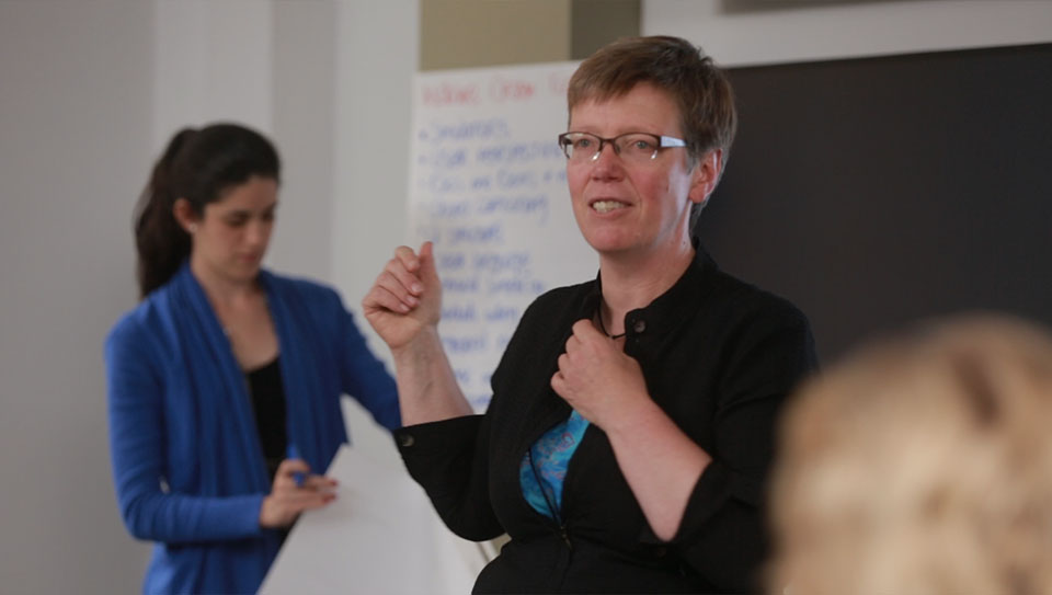 Two women address an audience and gesture at a poster of notes.